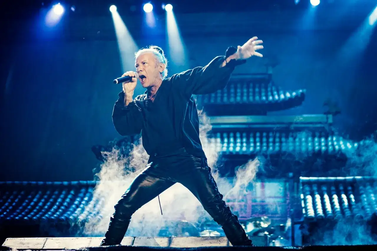 Bruce Dickinson, wokalista Iron Maiden, śpiewa na scenie otoczony dymem, w tle azjatycka scenografia.