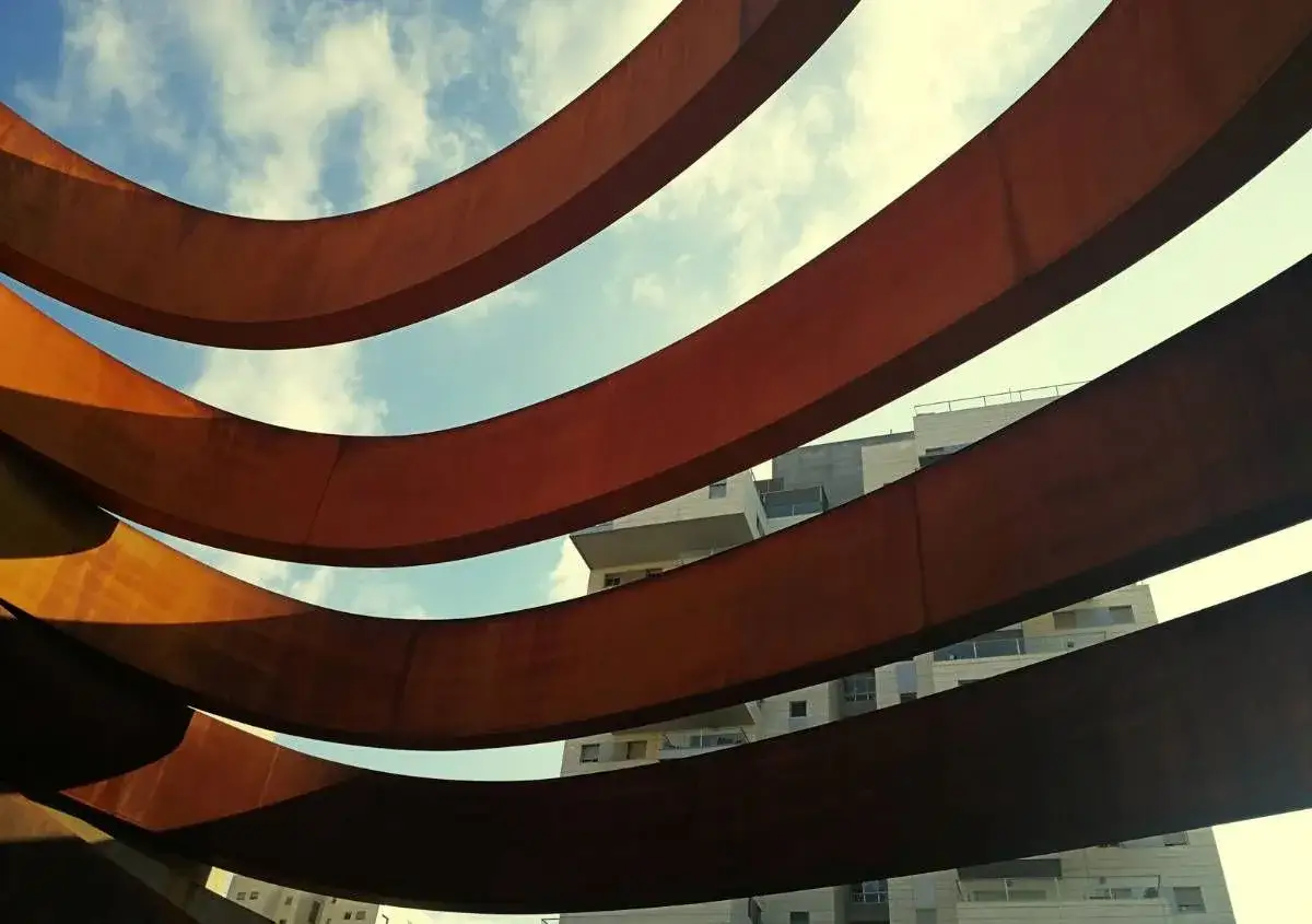 Struttura curva in corten materiale, con edifici moderni sullo sfondo e cielo azzurro con nuvole.
