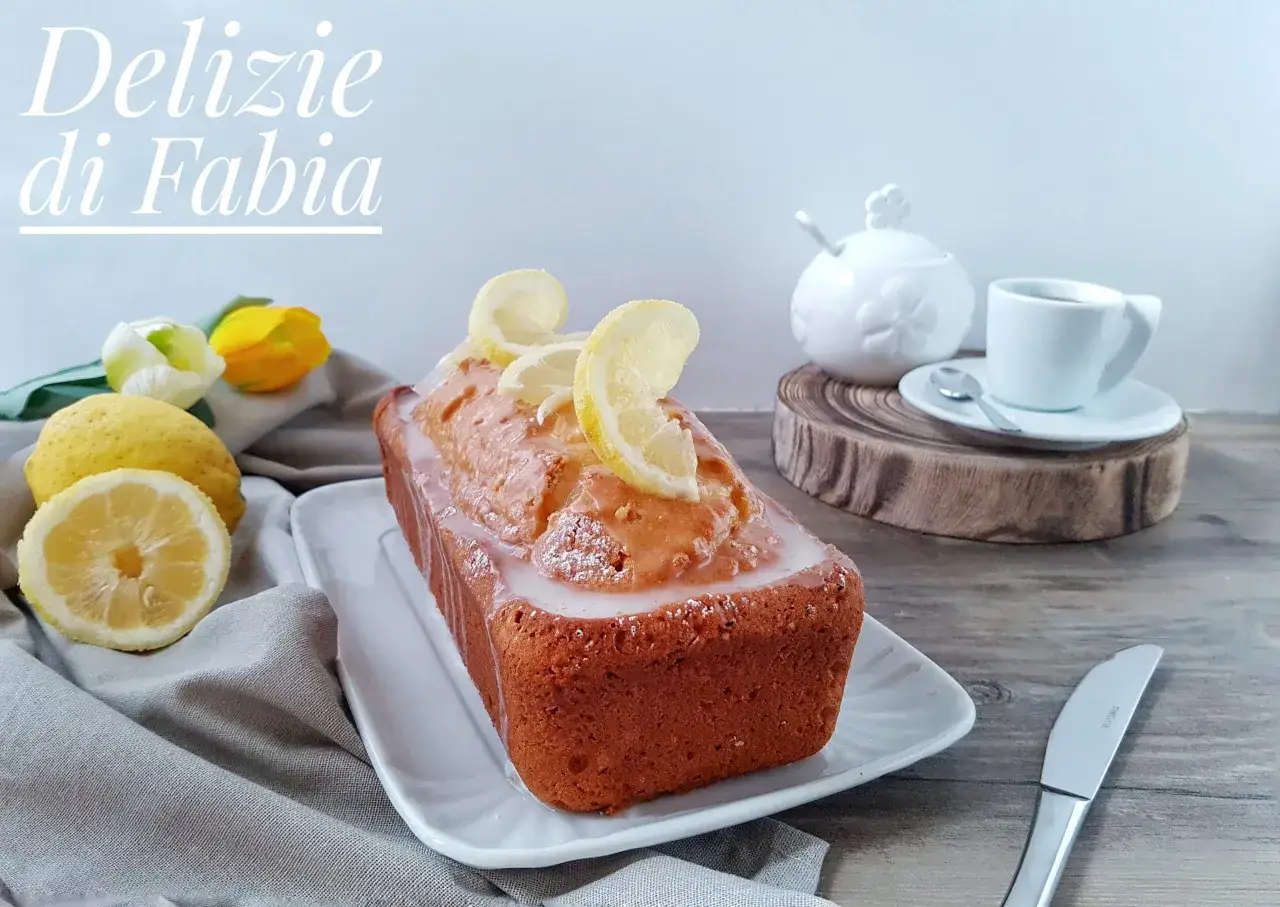 Delizioso plumcake al limone glassato, decorato con fettine di limone e spolverato di zucchero a velo. Accanto, limoni freschi e fiori.