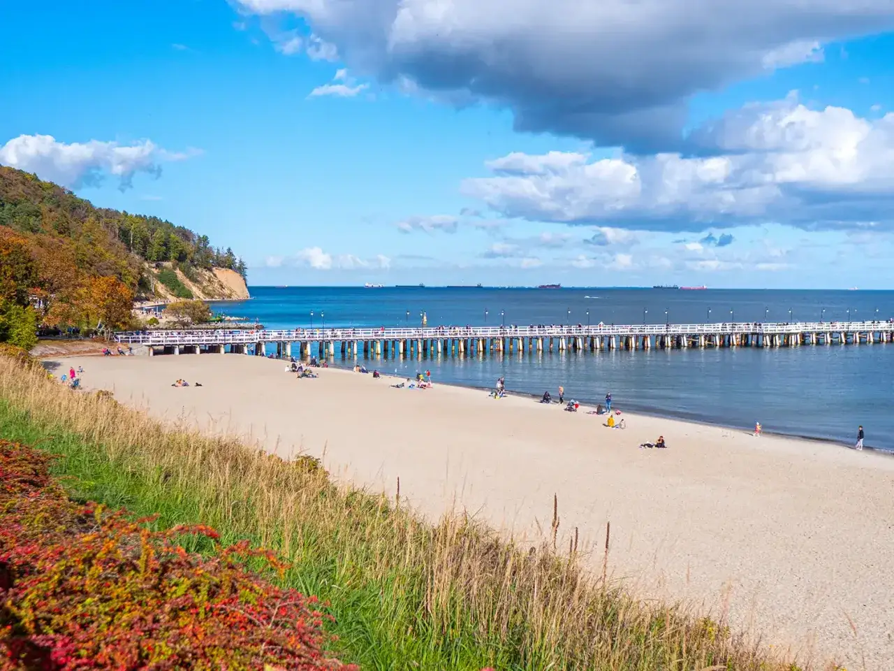 Słoneczna plaża w Sopocie z molo, ludźmi spacerującymi i odpoczywającymi na piasku. W tle widać klif i morze z żaglowcami.