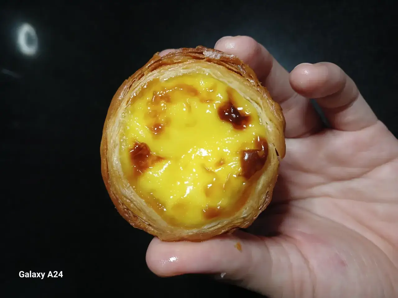 En hand h&aring;ller en gyllene, krispig pastel de nata med kr&auml;mig vaniljkr&auml;m och karamelliserade toppar.