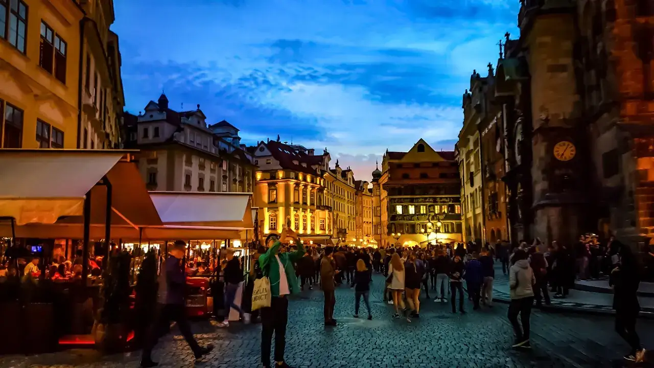 Abendliche Szene auf einem Platz in Prag. Menschen flanieren, Restaurants sind beleuchtet. Ein Ort, der an die lebhaften prag clubs erinnert.