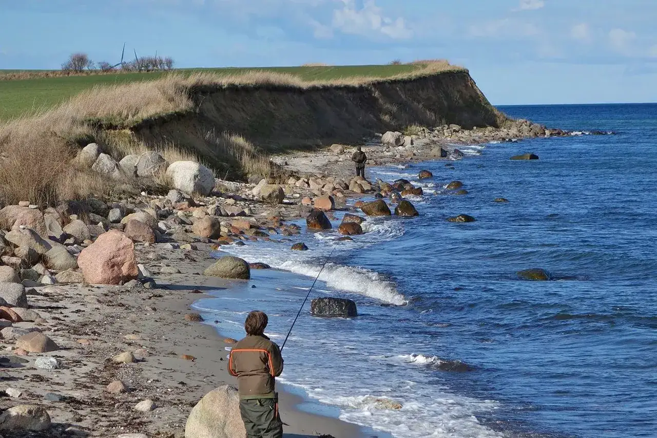 Ein Angler steht am steinigen Strand von Langeland und wirft seine Angel aus. Die Steilk&uuml;ste im Hintergrund und das blaue Meer sind aktuell ein idyllisches Bild.