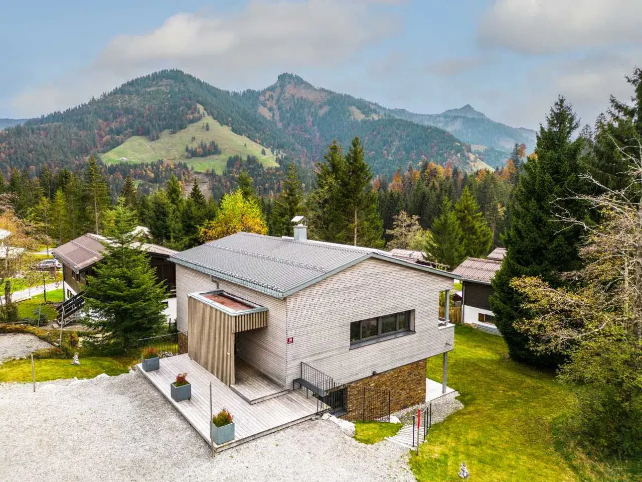 Modernes Haus in Hanglage mit Holzfassade, umgeben von Bäumen und Bergen im Herbst.