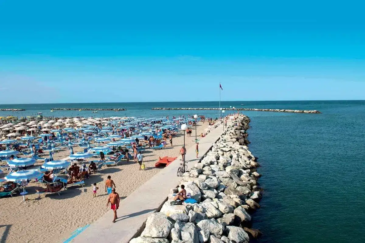Spiaggia affollata con ombrelloni e lettini a Rimini Gatteo Mare. Un molo in pietra si estende nel mare azzurro.