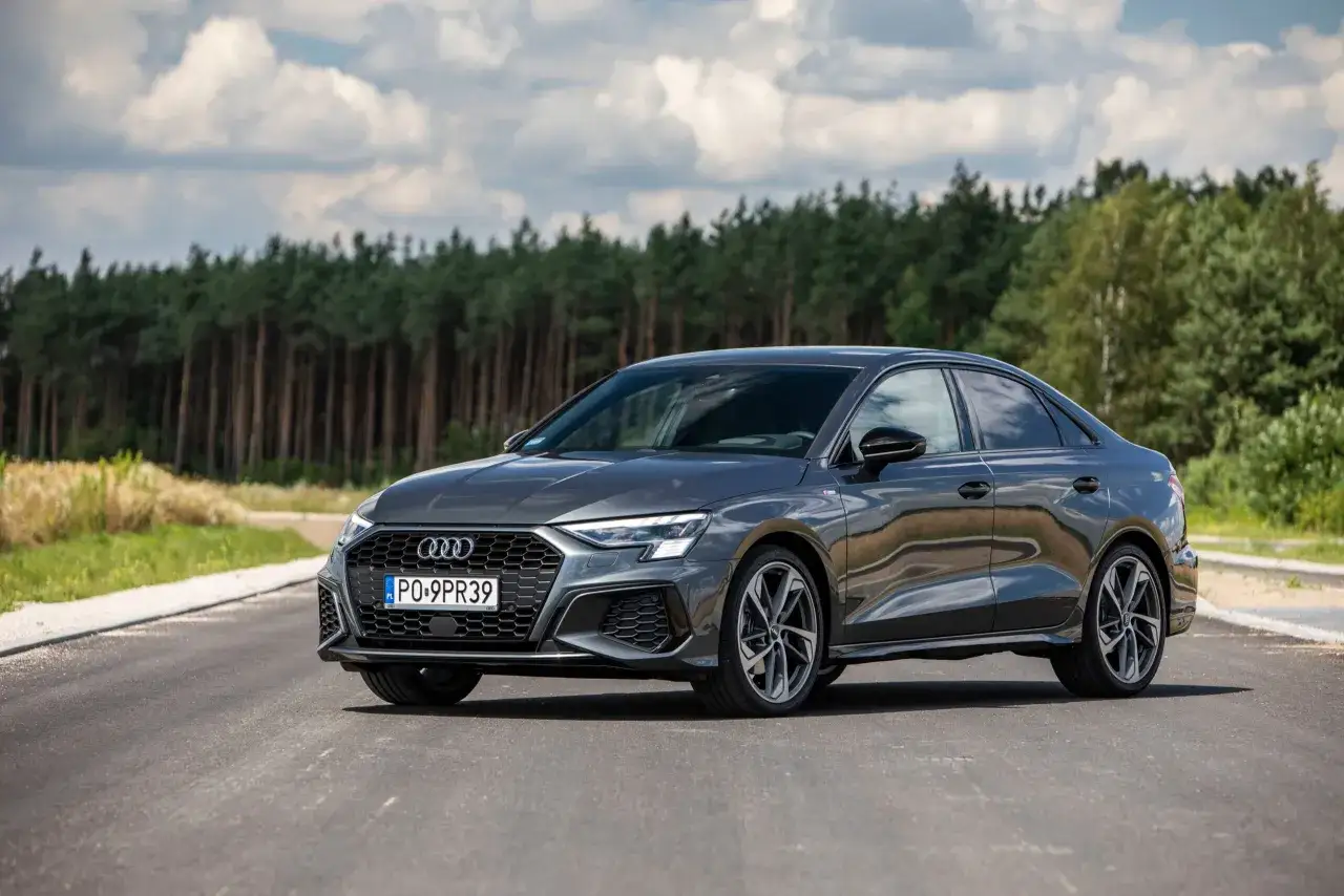 Szary sedan Audi A3, z grillem w kształcie plastra miodu i ostrymi światłami, zaparkowany na drodze z lasem w tle. Idealne wymiary a3 do prezentacji.