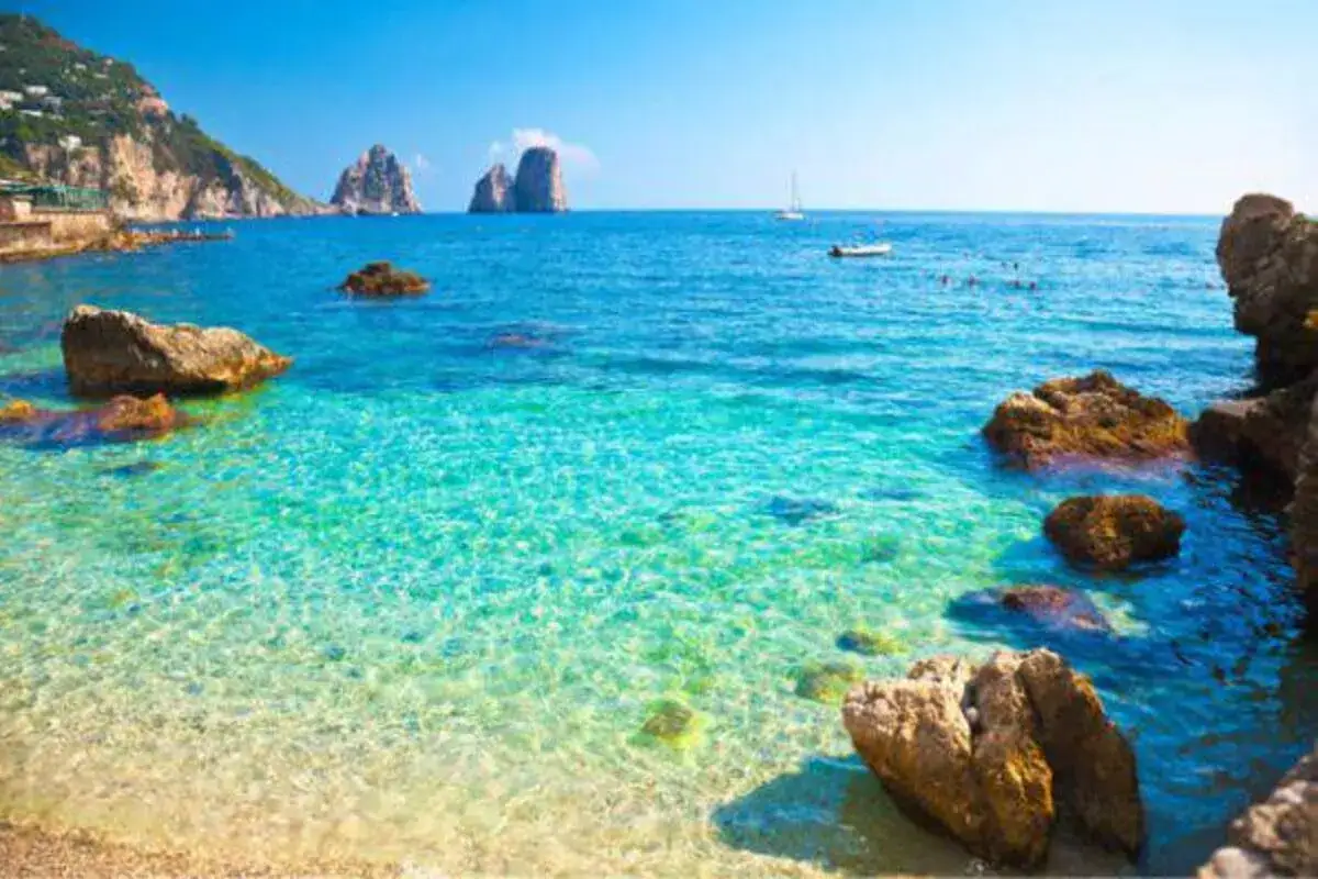 Acque cristalline e rocce a picco sul mare bello in Campania, con i Faraglioni di Capri all'orizzonte.