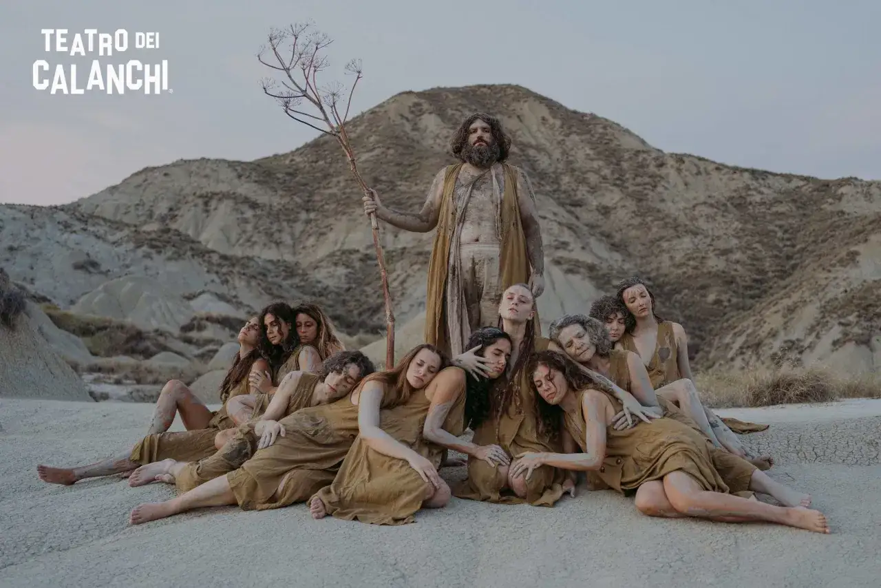 Un uomo barbuto con un ramo secco domina un gruppo di donne sdraiate, immerse nella terra. Un'evocazione del **teatro dei calanchi**.