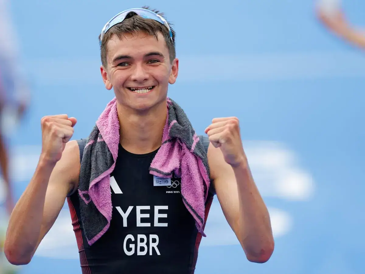 Alex Yee, triumphant and beaming, pumps his fists in celebration. A towel is draped around his neck, and his racing bib reads