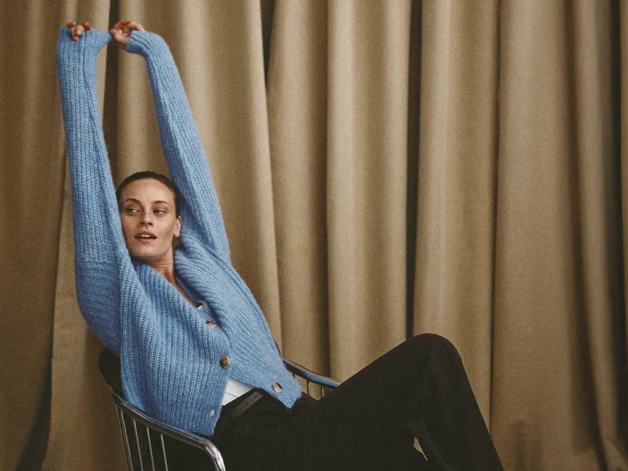 Kobieta w niebieskim swetrze z rozciągniętymi ramionami, siedząca na krześle. Stylowa, wygodna stylizacja.