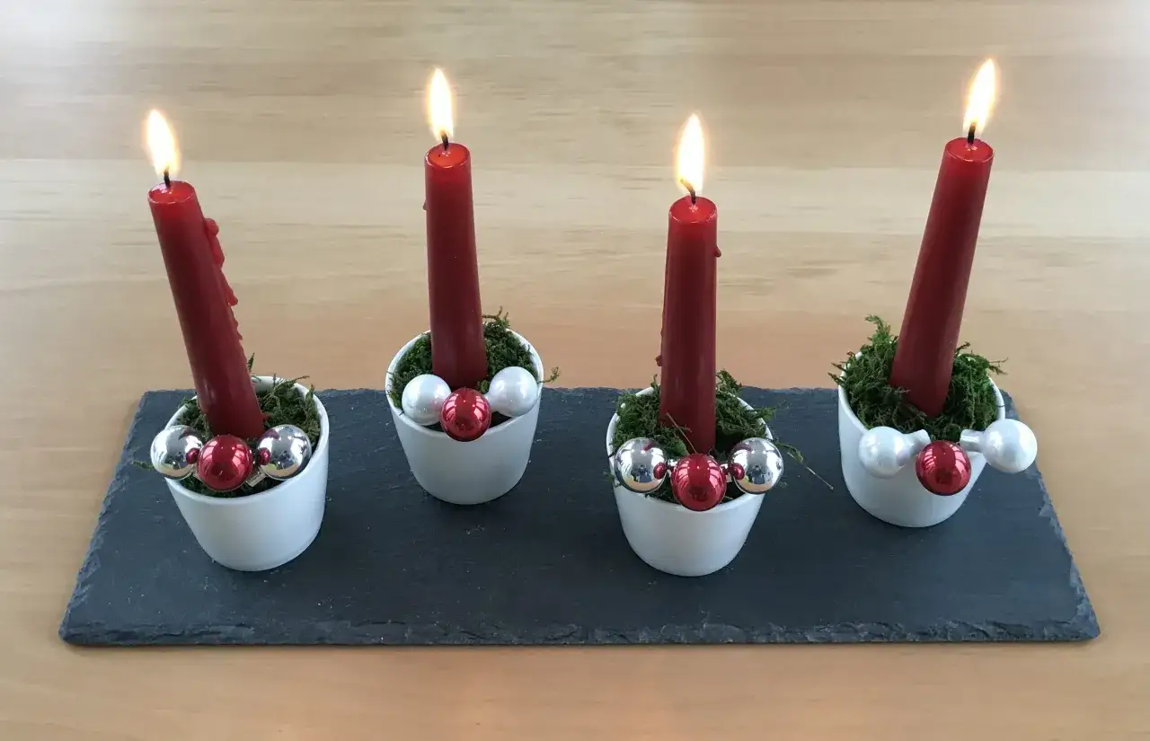 Vier rote Stabkerzen auf Moos in weißen Schalen, geschmückt mit Kugeln. Ein schöner Adventskranz, den man leicht selber machen kann.