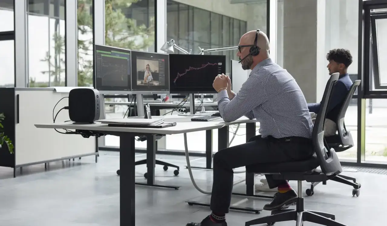 Men in an office using thin clients for government work. One man wears a headset, facing dual monitors displaying charts and a video call.