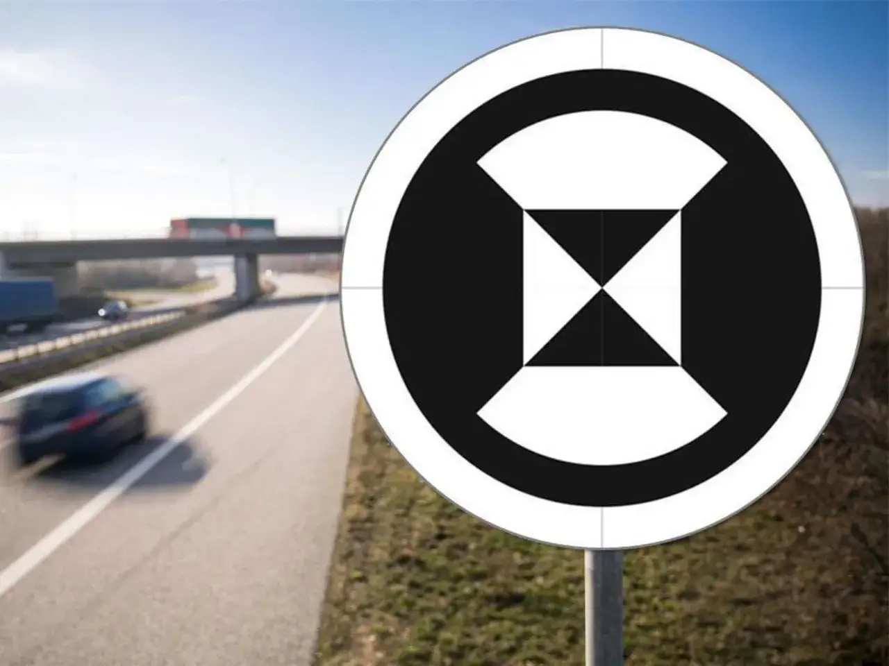 Ein rundes autobahn schild mit schwarzem Muster auf weißem Grund steht am Straßenrand. Im Hintergrund fährt ein Auto auf der Autobahn.