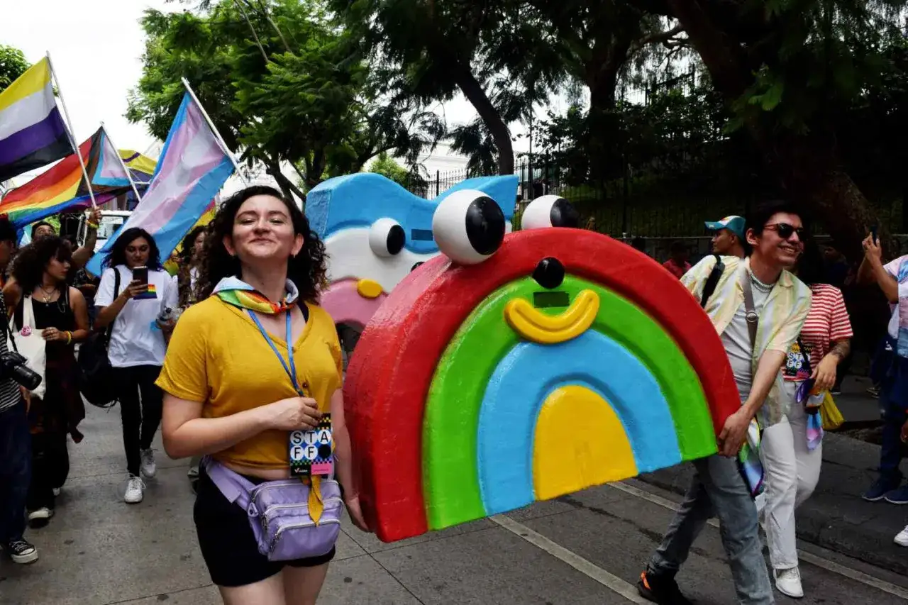 A vibrant parade celebrates gay pride in Guatemala. People carry rainbow flags and a large, colorful rainbow float.