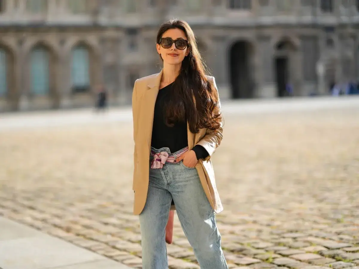 Kobieta w beżowej marynarce i jasnych mom jeans, zastanawia się jakie buty do mom jeans będą najlepsze.