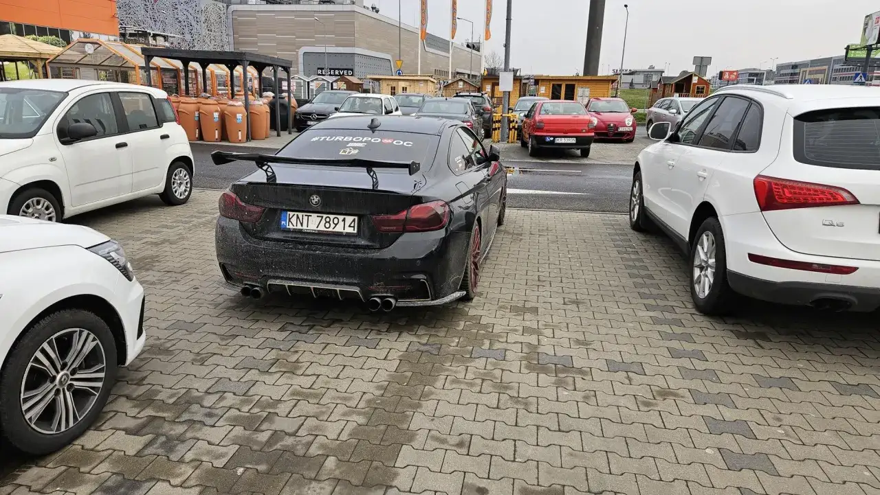 Czarny BMW z tablicą KNT 78914 i spoilerem na parkingu. Obok białe Audi i Fiat.
