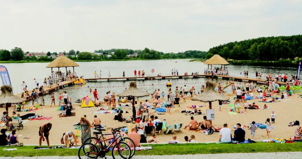 Tłum ludzi odpoczywa na plaży nad zalewem Siemiatycze. Woda, molo z altanami i zielone drzewa w tle.