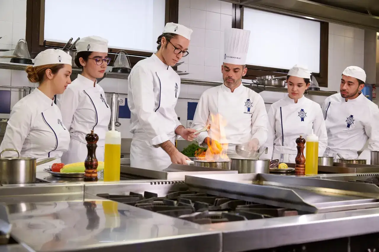 Estudiantes de **carrera de gastronom&iacute;a** aprenden t&eacute;cnicas culinarias en una cocina profesional, con un chef demostrando flambeado.