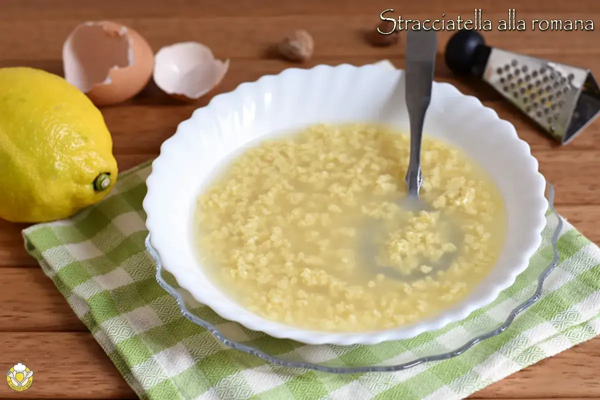 Stracciatella alla romana in una ciotola bianca, con limone, gusci d'uovo e grattugia sullo sfondo.