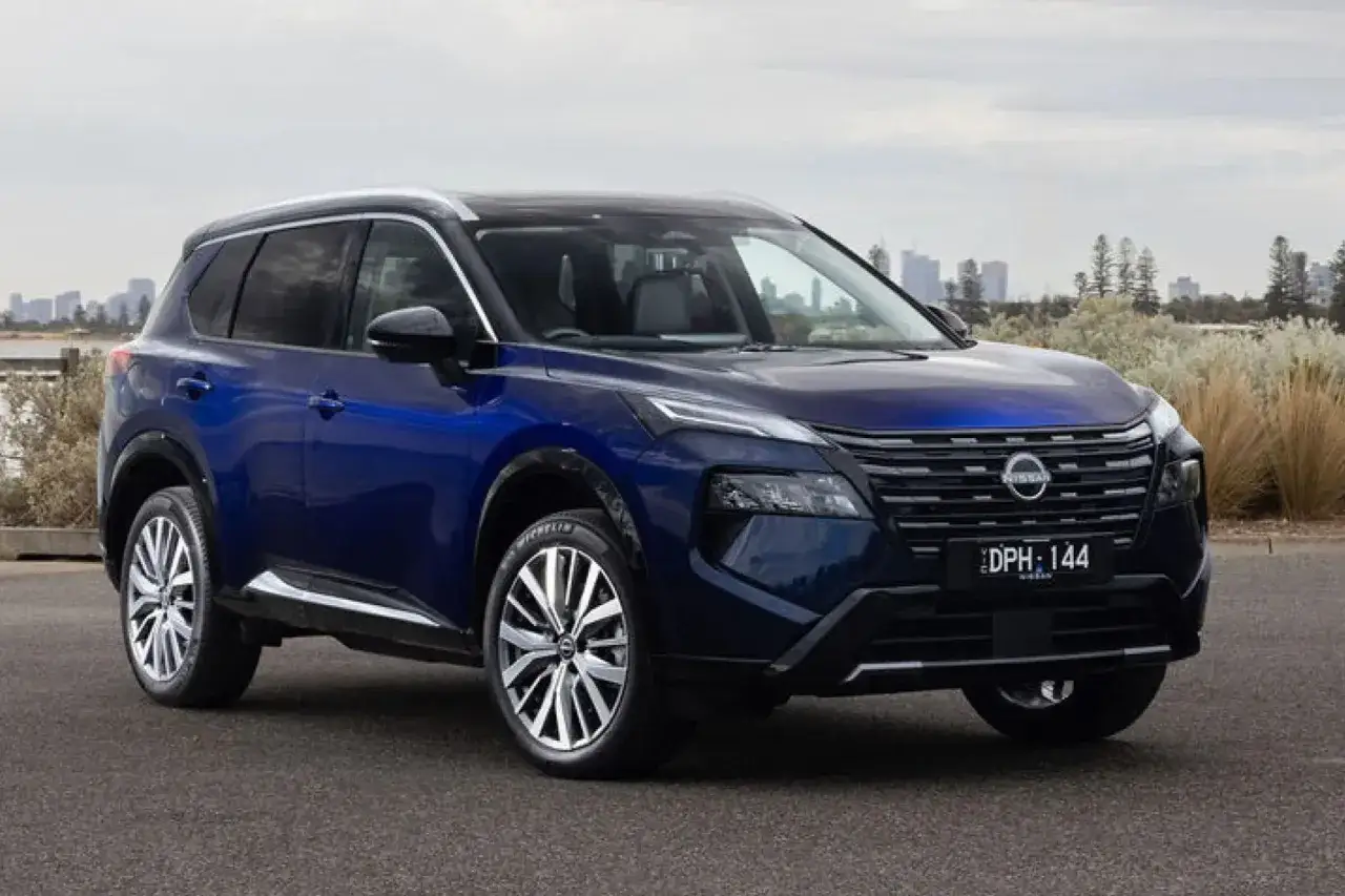 Bl&aring; Nissan X-Trail parkerad vid en strand med stad i bakgrunden. Inga synliga problem med bilen.