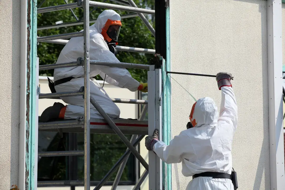 Deux ouvriers en combinaison intégrale et masque travaillent sur un échafaudage. Ils retirent des matériaux potentiellement dangereux. Amiante au travail, que faire ?