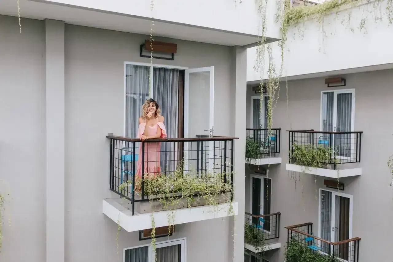 Kobieta w różowej sukience na balkonie, otoczonym zielenią. Rośliny pnące zwisające z góry sugerują, czym osłonić balkon, tworząc naturalną osłonę.