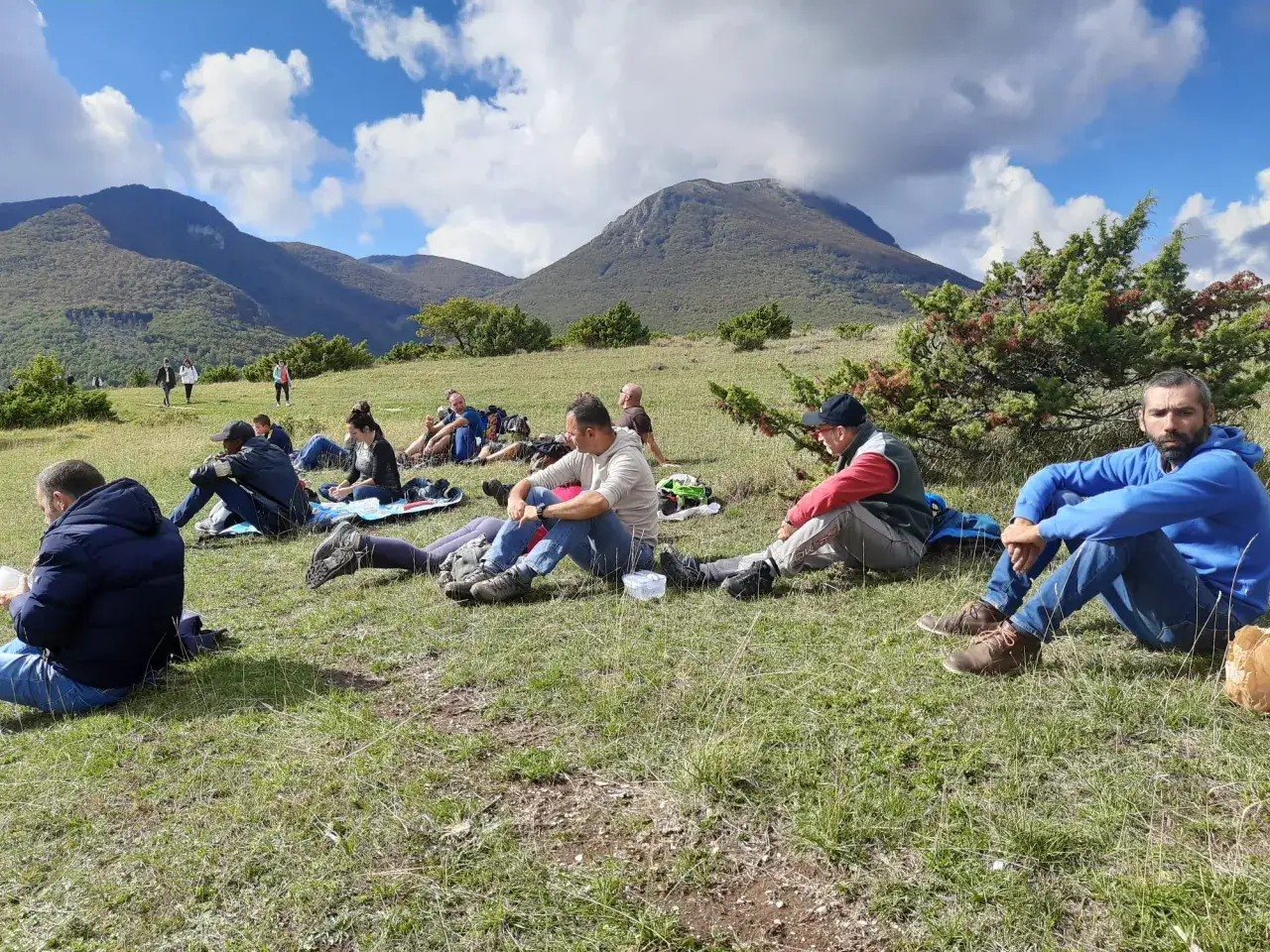 Gruppo di persone sedute sull'erba, con il monte San vicino sullo sfondo, sotto un cielo azzurro con nuvole bianche.