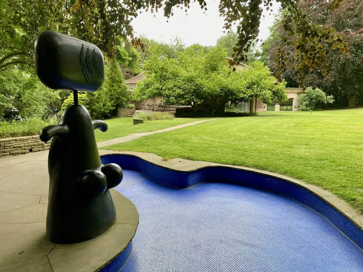 Eine abstrakte schwarze Skulptur steht am Rand eines blauen Mosaikbeckens in einem **Skulpturenpark**. Im Hintergrund eine gr&uuml;ne Rasenfl&auml;che und B&auml;ume.