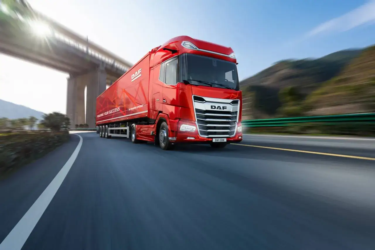 Un camion rouge DAF XF file sur une route, sous un pont, avec des montagnes en arri&egrave;re-plan.