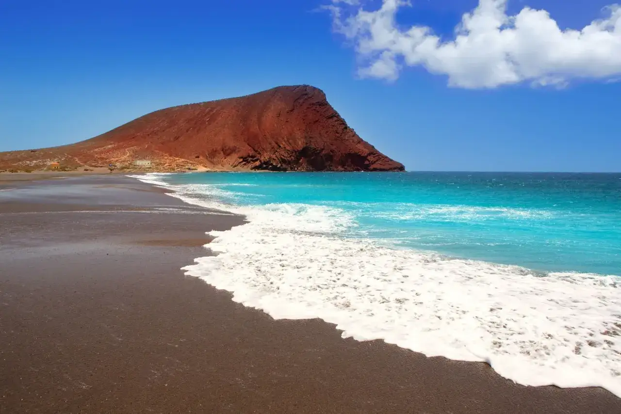 Plage de sable noir avec une montagne rouge et une mer turquoise. Idéal pour visiter Tenerife et profiter du soleil.