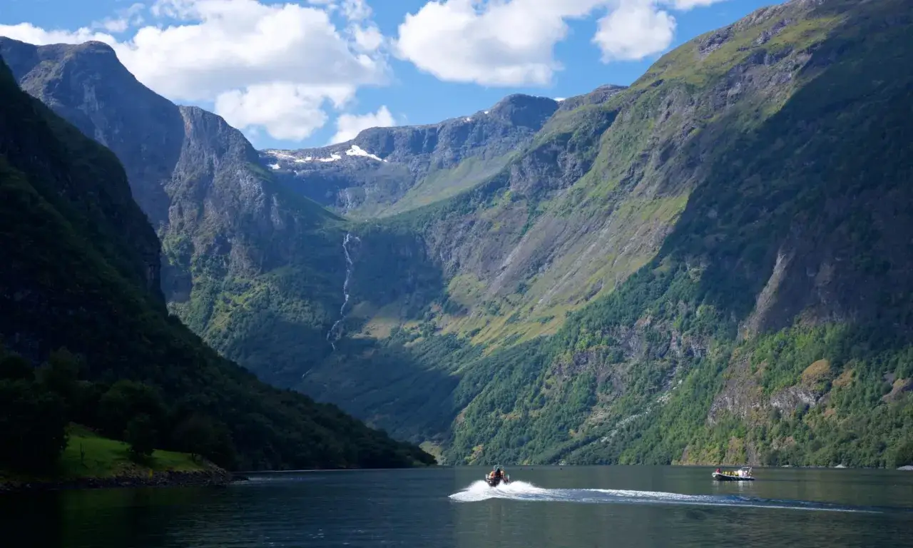 En båt susar fram på en fjord i Norge, omgiven av dramatiska berg och grönska. Kanske är de ute och fiska i norge.