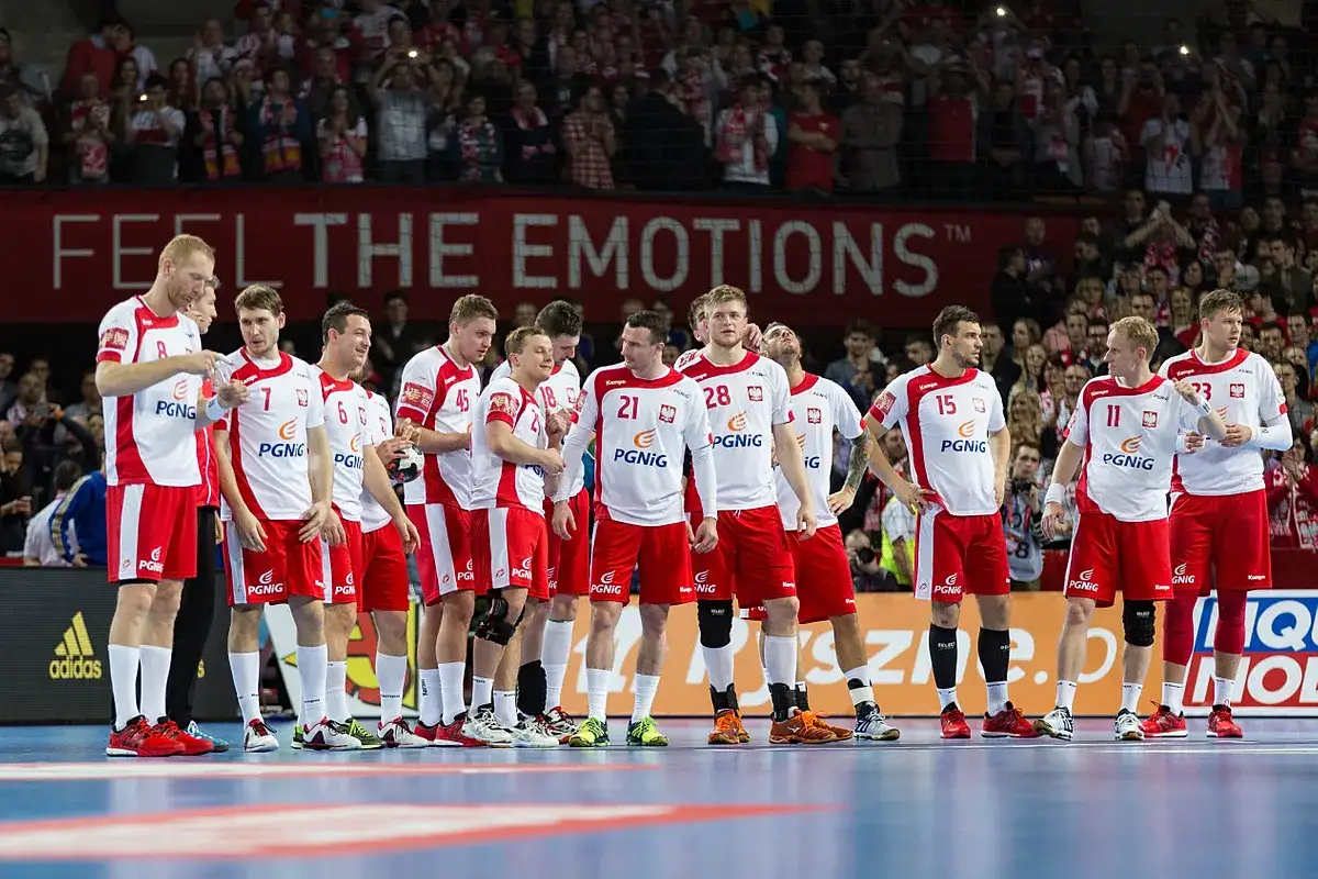 Drużyna piłkarzy ręcznych w białych strojach z czerwonymi elementami, w tym Bartosz Kuczyński, stoi na parkiecie przed trybunami pełnymi kibic&oacute;w.
