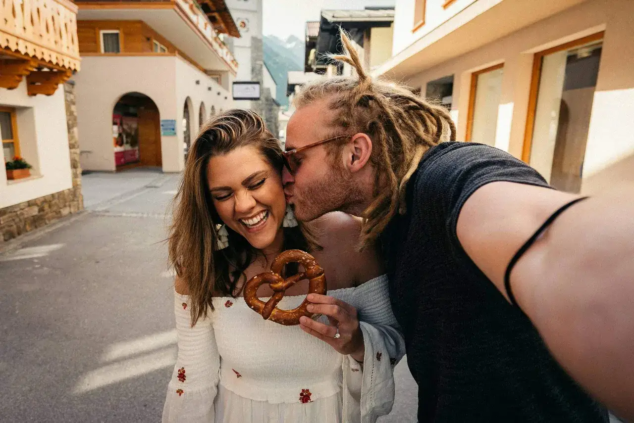 Ein Mann k&uuml;sst eine lachende Frau, die eine Brezel h&auml;lt. Ihre Freude zeigt Geduld in der Liebe, w&auml;hrend sie durch eine charmante Gasse schlendern.