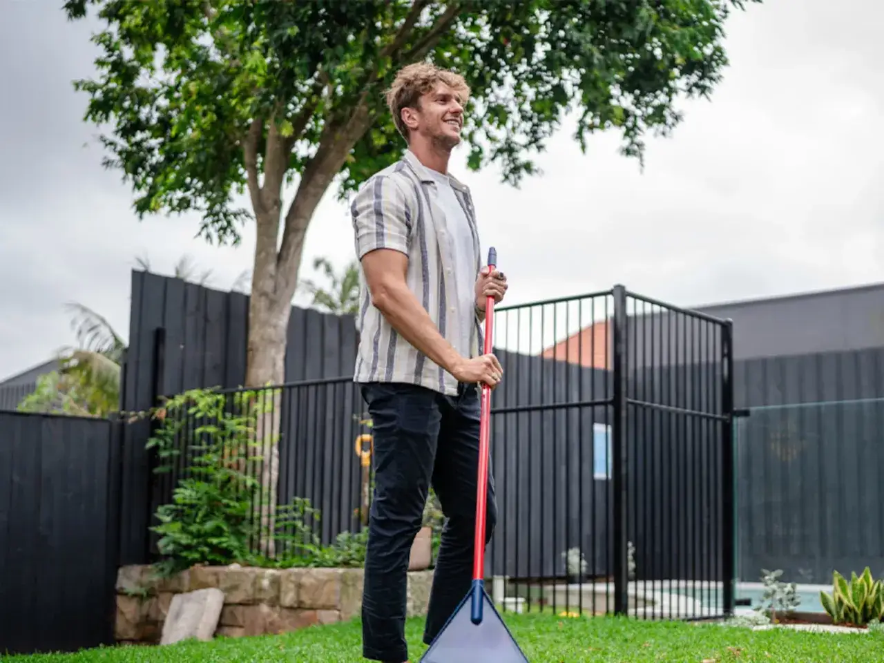 Hombre sonriente con rastrillo, listo para aprender como limpiar césped artificial y mantener su jardín impecable.