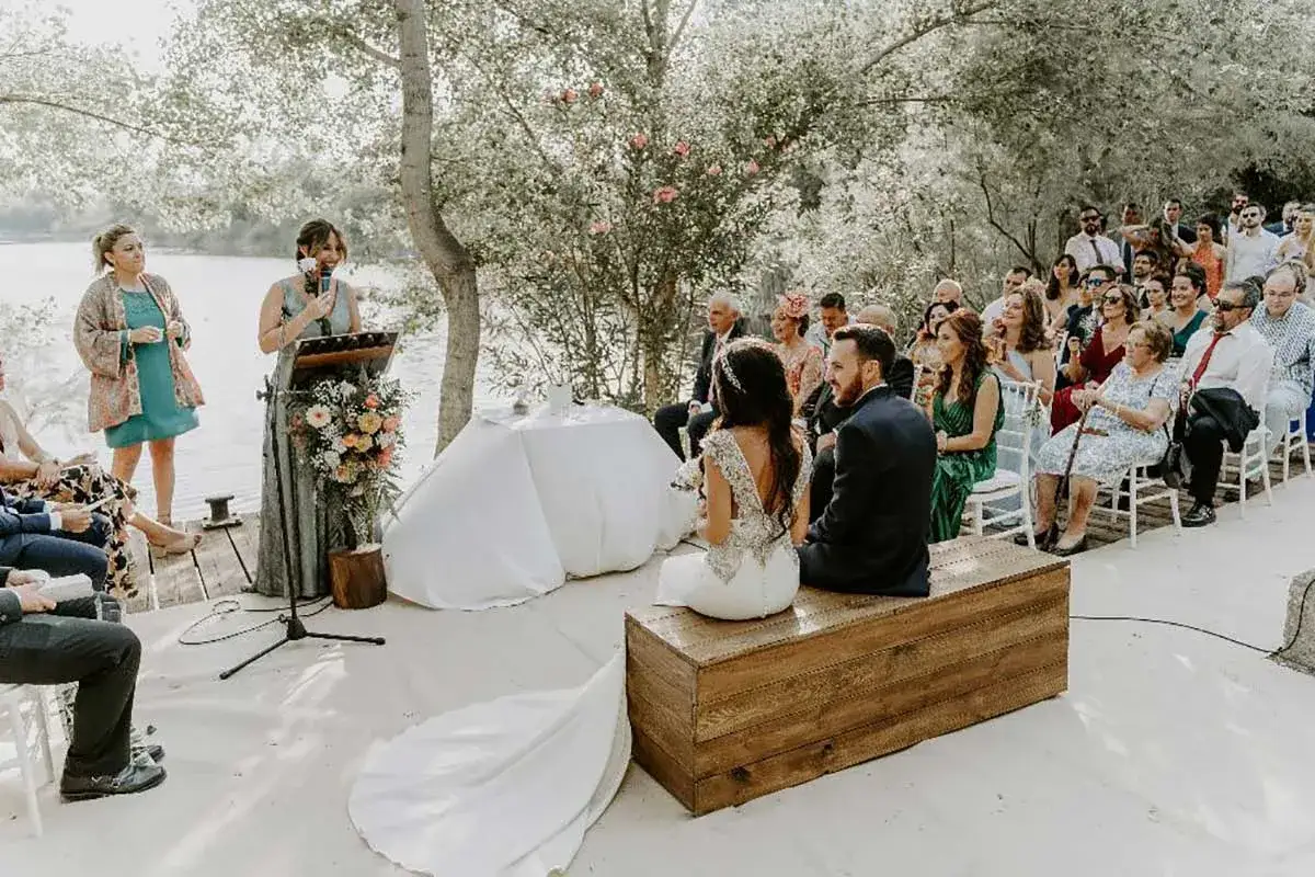 Una mujer lee textos para leer en una boda a los novios, sentados frente a invitados en una ceremonia al aire libre junto a un lago.
