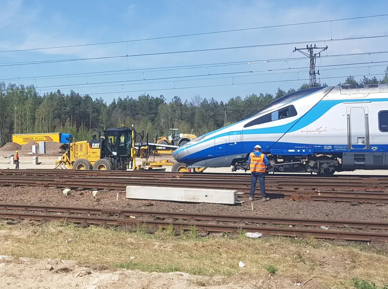 Prace torowe przy nowoczesnym pociągu Pendolino. W tle maszyny budowlane i las. To przyszłość prędkości pociągów w Polsce.