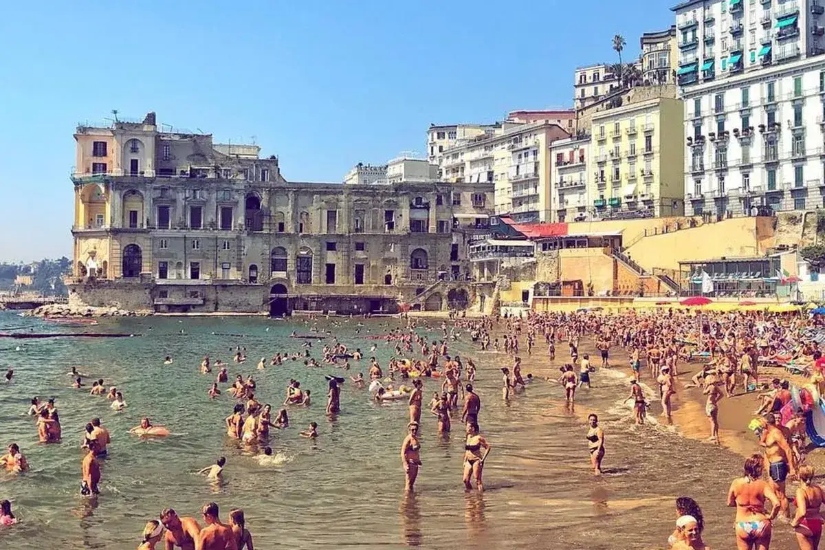 Un affollato scenario estivo sulle spiagge libere di Napoli, con bagnanti che si godono il mare e il sole davanti a palazzi storici.