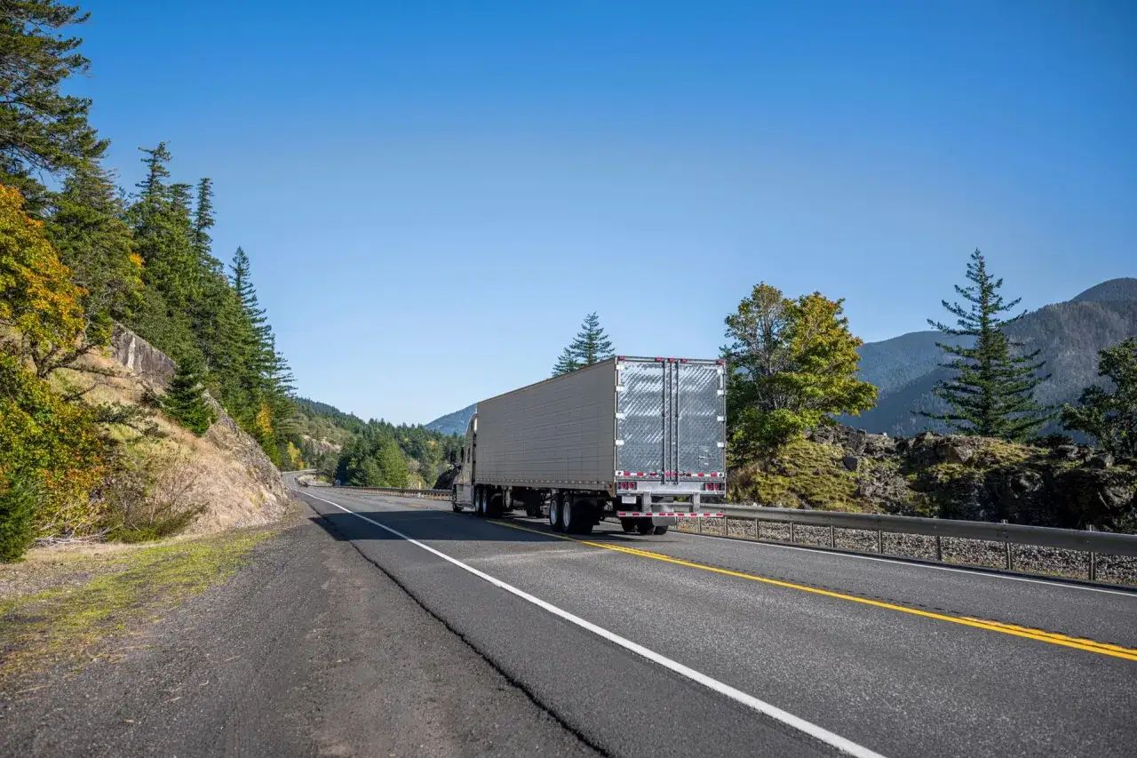 Un camionneur am&eacute;ricain traverse une route sinueuse bord&eacute;e d'arbres aux couleurs d'automne, sous un ciel bleu clair.