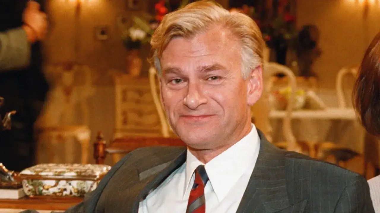 Peder Falk, en man med blont hår och en grårandig kostym, ler mot kameran. Bakgrunden är en elegant inredd salong.