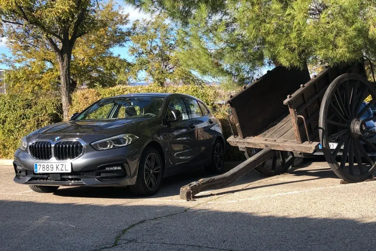 Un BMW Serie 1 116d gris aparcado junto a un carro de madera antiguo. Parece que el coche tiene algunos problemas, quizás relacionados con el motor o la transmisión.