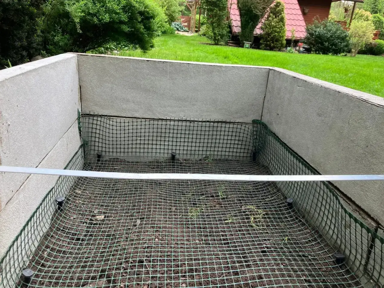 Betonowa konstrukcja z siatką zabezpieczającą przed kretami. W tle zielony trawnik i domki.