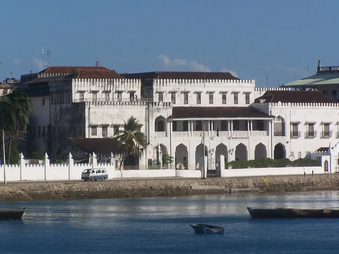 Biały pałac z arkadami i palmami, symbol życia na Zanzibarze. W oddali widać łodzie na wodzie.