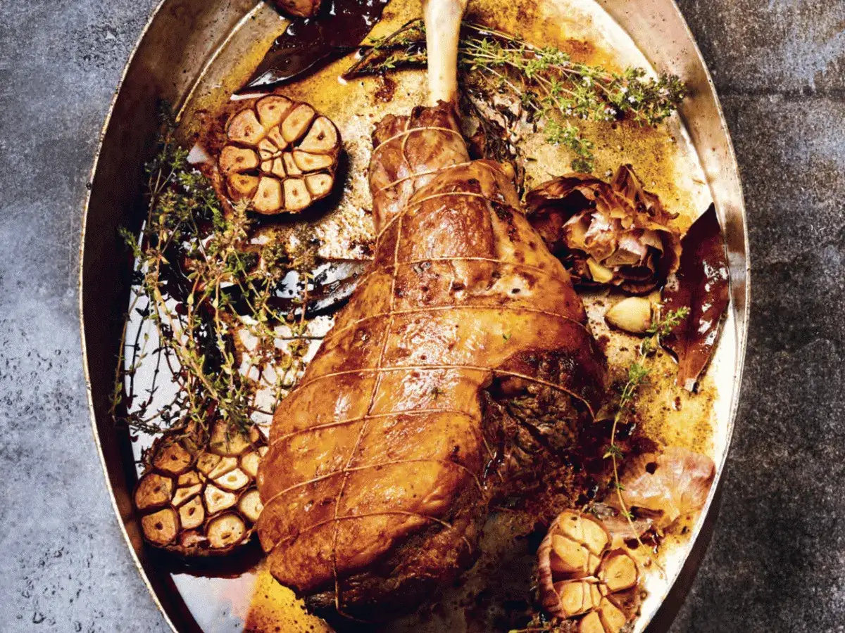 Gigot d'agneau r&ocirc;ti pour 6 personnes, servi dans un plat avec ail confit, thym et laurier.