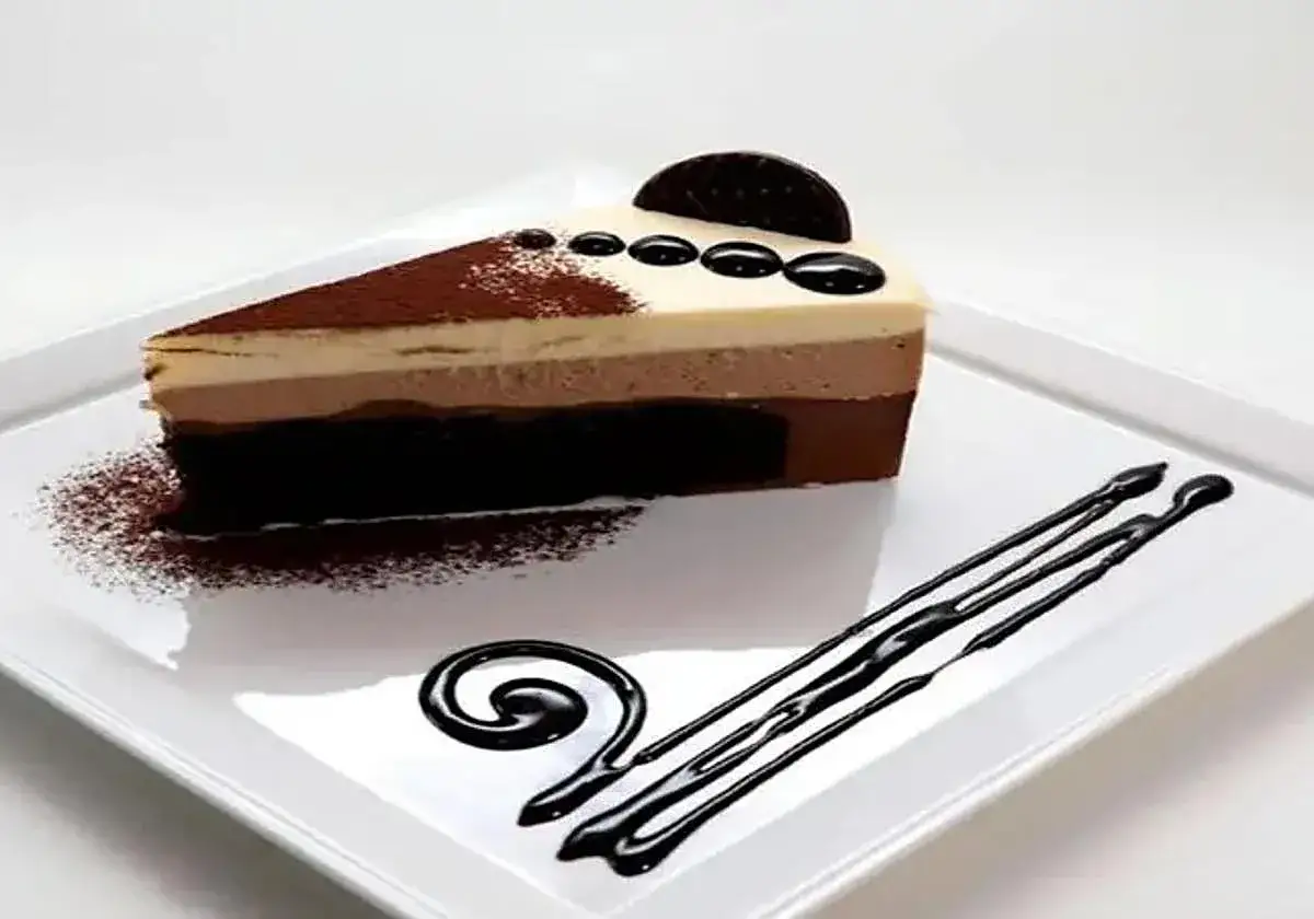 Una rebanada de tarta de tres chocolates, con capas de mousse de chocolate blanco, con leche y negro, espolvoreada con cacao y decorada con chocolate.