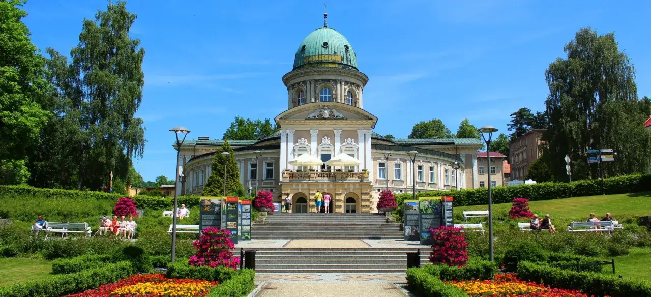 Elegancki budynek z zieloną kopułą, otoczony kwitnącym ogrodem i drzewami. W oddali widać ludzi odpoczywających na ławkach. To idealne miejsce na relaks i ładowanie baterii.
