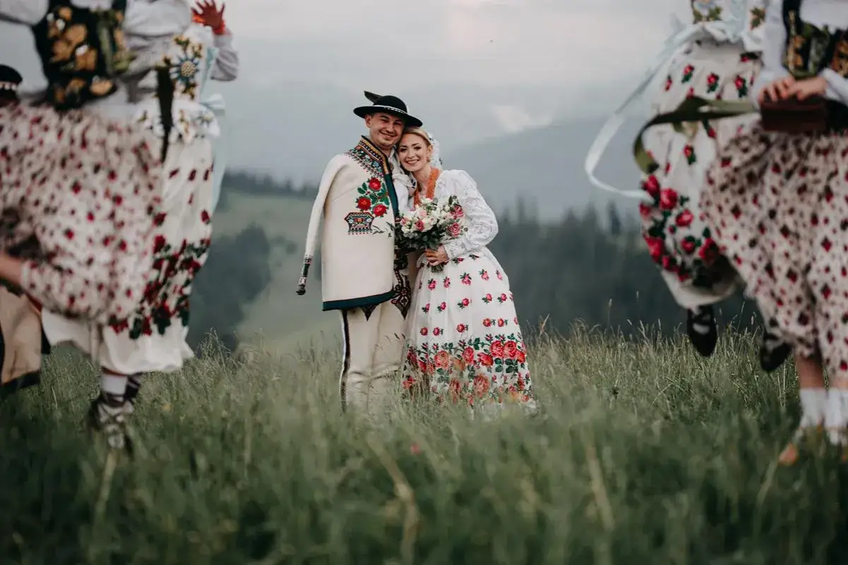 Para młoda w strojach ludowych, otoczona tańczącymi kobietami w tradycyjnych strojach, na tle gór. Słychać radosne przyśpiewki góralskie.