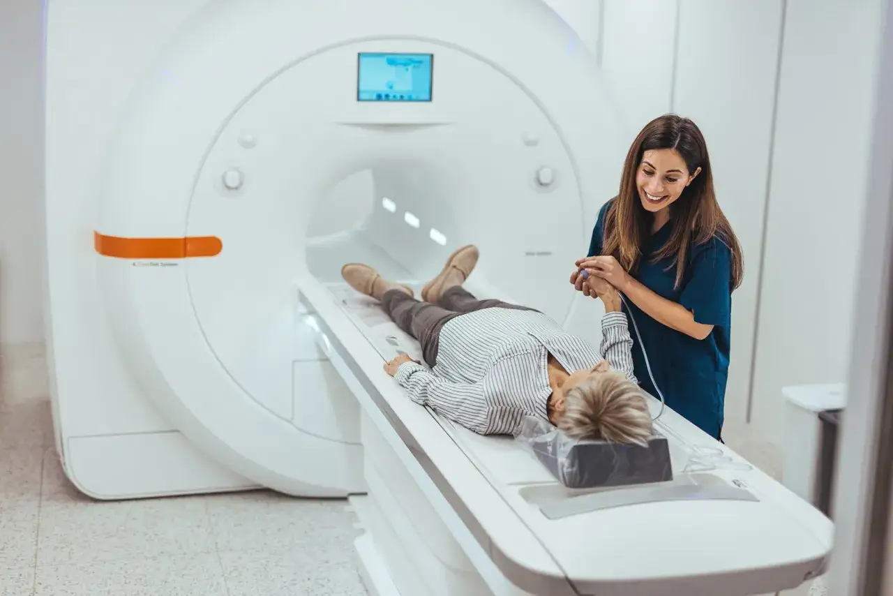 Eine Patientin liegt f&uuml;r eine MRT Untersuchung auf einer Liege, w&auml;hrend eine freundliche &Auml;rztin ihr die Hand h&auml;lt.
