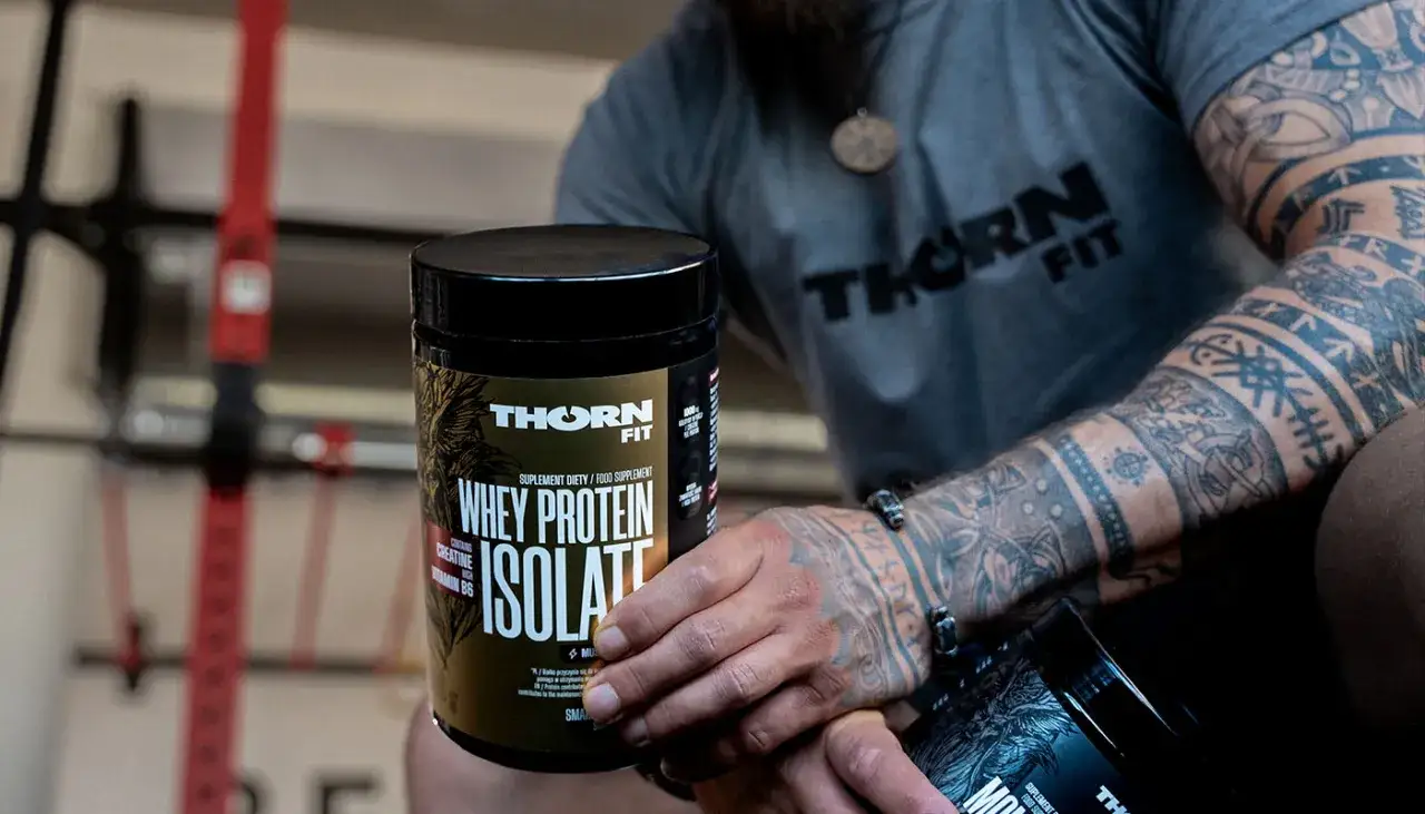 Mężczyzna z tatuażami trzyma opakowanie odżywki białkowej Thorn Fit. Idealne białko po treningu dla regeneracji mięśni.