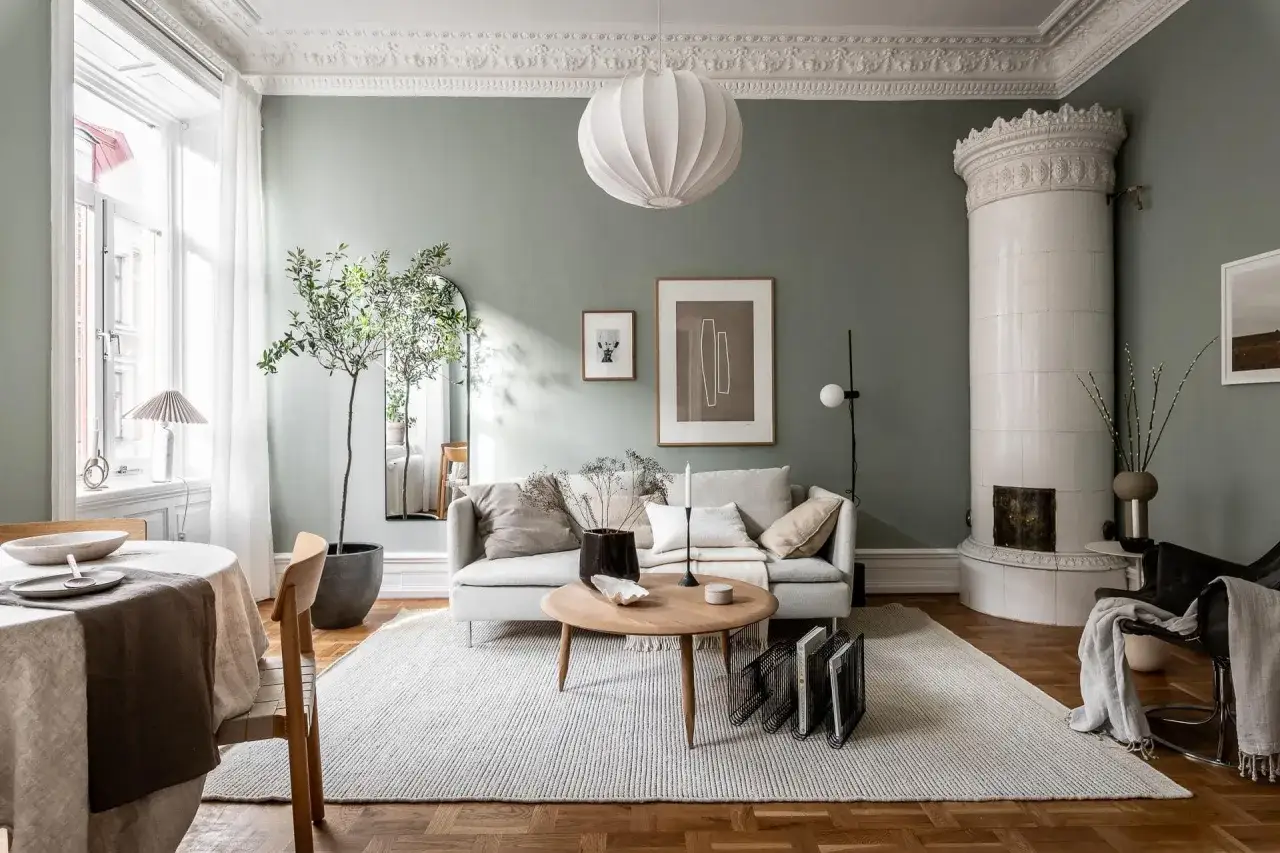 Salon avec murs vert sauge et sol en parquet. Un canap&eacute; gris clair est plac&eacute; sur un tapis blanc. Quelle couleur de mur avec un sol gris fonc&eacute; ? Ce salon est une inspiration.