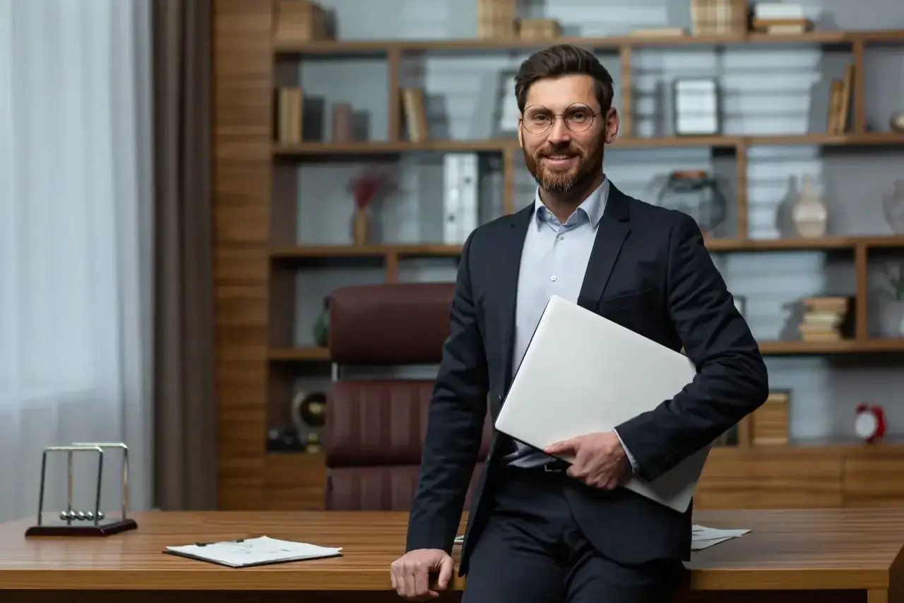 Uśmiechnięty dyrektor szkoły podstawowej w garniturze, trzymający laptopa, stoi w swoim gabinecie.